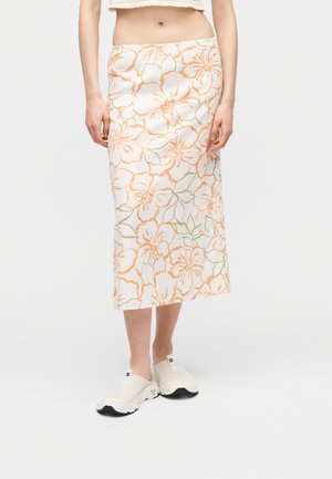 Kvinde iført en hvid midinederdel med orange blomsterprint og grønne blade, kombineret med hvide slip-on sneakers, stående mod en ensfarvet baggrund.