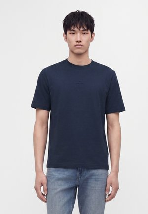 Jeune homme aux cheveux courts noirs porte un T-shirt bleu marine uni foncé et un jean bleu clair, debout devant un fond blanc.