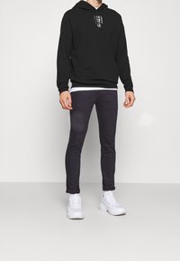 Sudadera negra con capucha y bolsillo frontal, camiseta blanca debajo, combinada con jeans ajustados gris oscuro y zapatillas blancas.