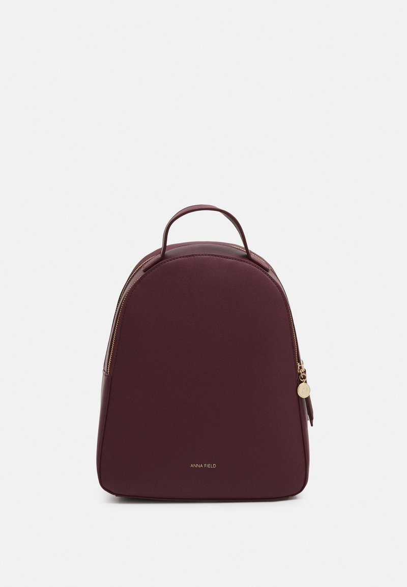 Sac à dos compact couleur bordeaux avec une poignée supérieure, des détails de fermeture éclair dorée et un petit logo "Anna Field" au centre avant.