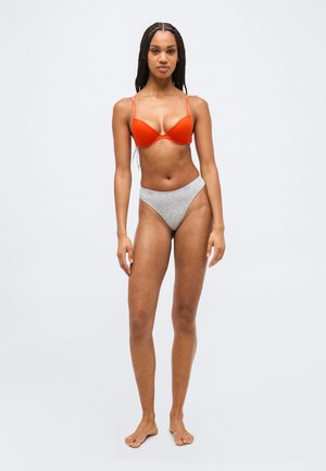 Γυναίκα όρθια ξυπόλητη, φοράει φωτεινό πορτοκαλί σουτιέν και ανοιχτό γκρι εσώρουχο με λευκή μέση, σε απλό φωτεινό φόντο.