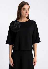 Czarny krótki top z rękawami i okrągłym dekoltem. Posiada duży tkaninowy akcent w kształcie serca po lewej stronie, dodający faktury. Gładki materiał.