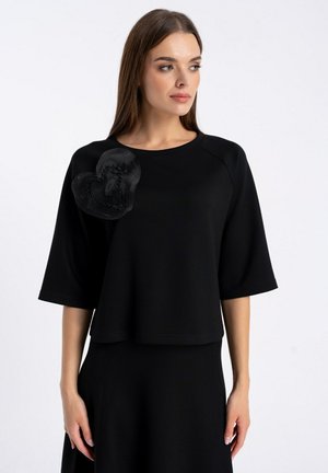 Czarny krótki top z rękawami i okrągłym dekoltem. Posiada duży tkaninowy akcent w kształcie serca po lewej stronie, dodający faktury. Gładki materiał.