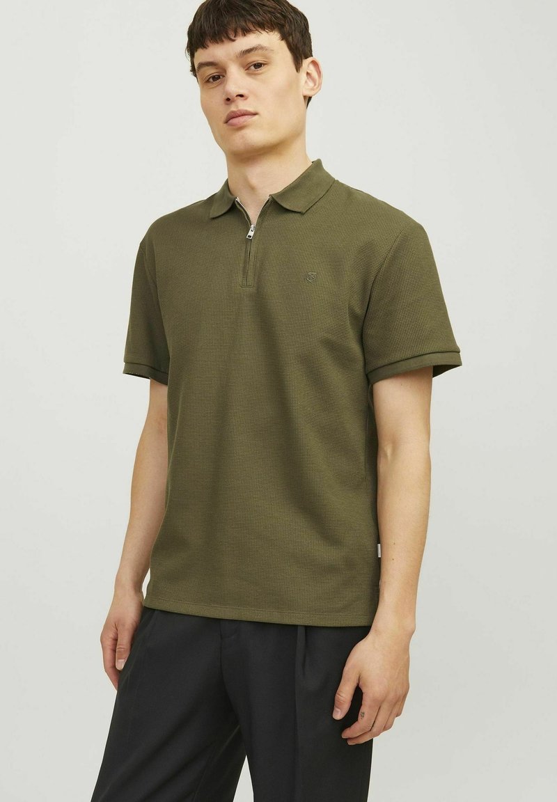 Olivgrön polo shirt med dragkedja i kragen, strukturerad tyg, korta ärmar och en subtil logotyp på bröstet. Bärs med mörka byxor.