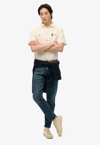 Camicia beige a maniche corte con bottoni, jeans scuri e sneakers beige chiare, completata da un maglione blu navy legato in vita e un piccolo dettaglio con patch.