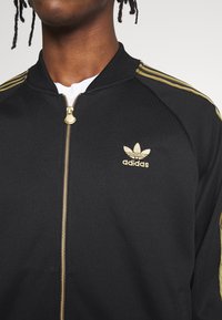 Svart Adidas zip-jacka med guldstripes och logga. Har en texturerad tygkvalitet och guld dragkedja. Designen är sportig och figurnära.