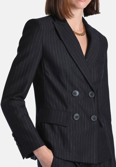 Femme portant un blazer noir à fines rayures, croisé à quatre boutons, avec un collier chaîne en or, main dans la poche.
