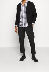 Cardigan nero sopra una camicia a scacchi blu e bianca con bottoni, abbinato a pantaloni neri e stivali neri con lacci, che mostrano un look a strati.
