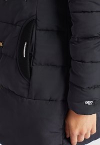 Detailní záběr na ruku osoby blízko kapsy černé polstrované bundy s viditelnými logy KHJO na manžetě a obvodu kapsy.