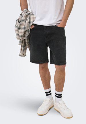 Mann iført hvite joggesko, hvite sokker med svarte striper, svarte denimshorts og en hvit T-skjorte, holder en beige rutete skjorte.