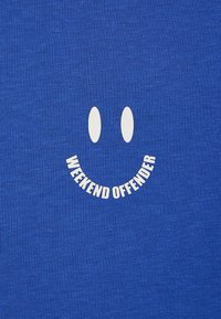 Zila kokvilnas T-krekls ar baltu smaidoša sejas grafiku, kurai zem acīm ir izliekts teksts "WEEKEND OFFENDER". Mīksta, teksturēta auduma.