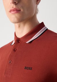 Crvena polo majica s teksturiranom tkaninom, dugmadi na prednjem dijelu i kontrastnim ovratnikom s crnom prugom. Izvezeni „BOSS“ logo u crnoj boji.