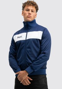 Junger Mann mit lockigem Haar, der eine navyblaue und weiße Lonsdale-Jacke mit Reißverschluss trägt, steht mit gefalteten Händen vor sich.