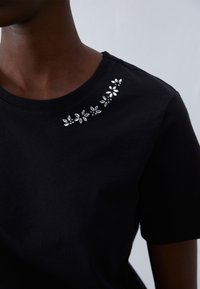T-shirt en coton noir avec un col rond et des embellissements floraux argentés sur la zone des épaules. Texture lisse.