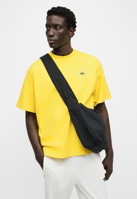 Άνδρας που φοράει κίτρινο T-shirt Adidas και άσπρο παντελόνι με μαύρη τσάντα χιαστί, στέκεται με τα χέρια στις τσέπες, κοιτάζοντας προς τα αριστερά.