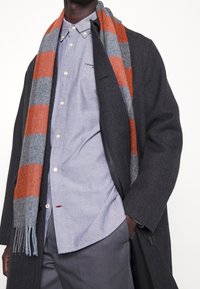 Harmaa villatakki sinisen napitettava paidan päällä, pariteltuna harmaasävyisen ja oranssin raidallisen huivin kanssa, jossa on hapsupäät.