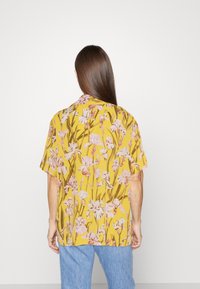 Kurzärmlige, gelbe Bluse mit floralem Muster, das pinke Blumen und grüne Stängel zeigt. Die Rückansicht zeigt einen lockeren Schnitt und ein Kragen-Design.