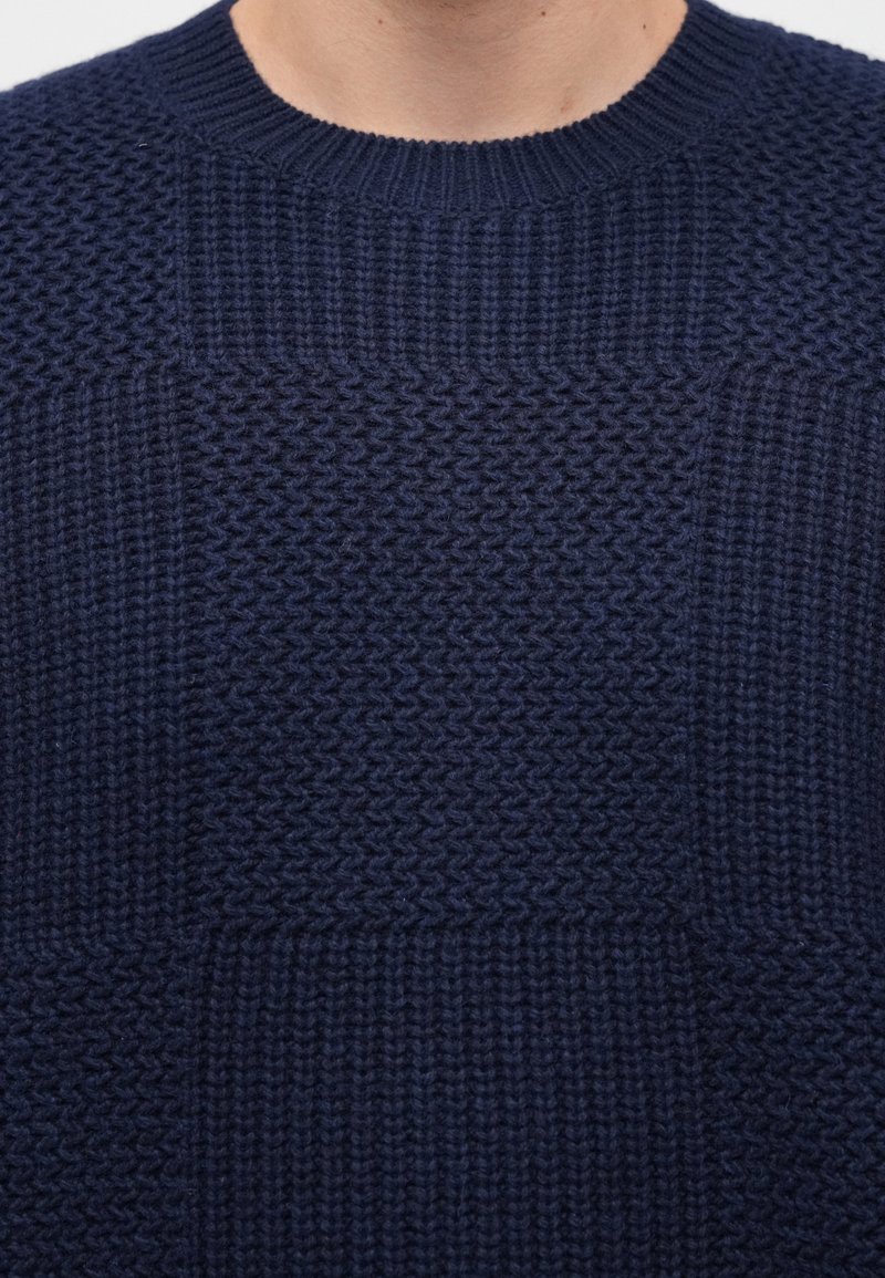 Navy knitted sweater featuring textured patterns and a ribbed neckline. The design includes square sections with varied knit patterns.