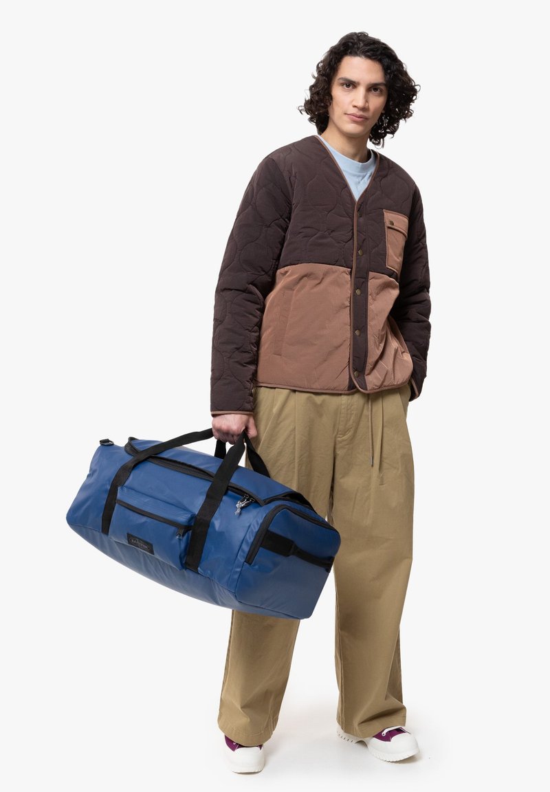Ge quiltete Jacke in Braun und Beige mit Knopfverschluss und Brusttasche, kombiniert mit übergroßen beigen Hosen. Blaue Duffle-Tasche mit schwarzen Akzenten.