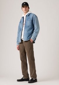 Giacca di jeans azzurro chiaro, felpa bianca con testo rosso, pantaloni in corduroy marroni e scarpe nere. Il modello indossa un cappellino nero con logo.