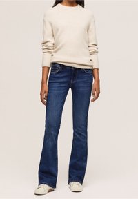 Beżowy sweter dzianinowy z prążkowanymi mankietami, w zestawieniu z ciemnoniebieskimi dżinsami dzwonami i białymi sneakersami. Dżinsy mają klasyczny pięciopocketowy krój.