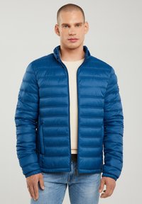 Mann trägt eine blaue gesteppte Jacke über einem weißen strukturierten Pullover und hellblauen Jeans, steht vor einem schlichten Hintergrund.