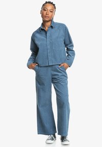 Cropped blue corduroy shirt with buttons, front pocket; matching wide-leg trousers. Worn with black and white sneakers.