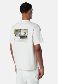 Hombre vistiendo una camiseta blanca con un gráfico en blanco y negro de un snowboarder y el texto amarillo "NORTH" en la parte de atrás, combinado con pantalones blancos.