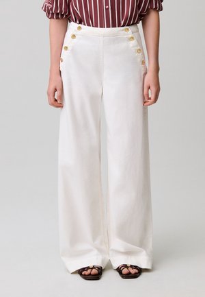 Pantalon blanc à jambes larges avec des boutons dorés sur les poches avant, porté avec des sandales marron à brides et une chemise bordeaux rayée aux manches retroussées.