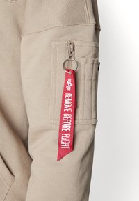 Manica di felpa beige con tasca con zip, dotata di un'etichetta rossa "RIMUOVERE PRIMA DEL VOLO". Il tessuto ha un aspetto texturizzato.