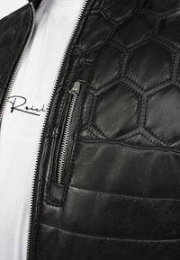 Zwart leren jack met een gewatteerd honingraatpatroon op de schouders, een ritszak op de borst en zilveren ritssluiting. Witte T-shirt zichtbaar eronder.