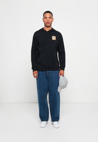 Felpa nera con cappuccio, tasca frontale e patch con logo, abbinata a jeans in denim blu larghi e un cappello grigio chiaro, con sneaker bianche.