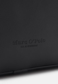 Surface en cuir noir embossé avec le texte "Marc O'Polo" et "Est. à Stockholm" en police plus petite en dessous.