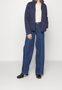 Granatowa pikowana kurtka z wysokim kołnierzem, w połączeniu z jasnobeżową bluzką i szerokimi dżinsami w kolorze niebieskim. Czarny botki na kostkę dopełniają stylizację.