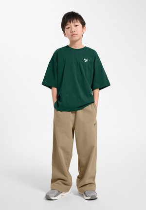 Chłopiec stoi z rękami w kieszeniach, ubrany w ciemnozielony oversize'owy t-shirt, beżowe szerokie spodnie i szare trampki na jednolitym tle.