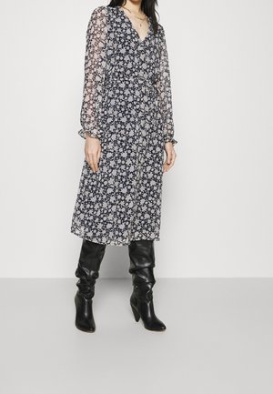 Vrouw die een zwart-witte midi-jurk met bloemenprint en doorschijnende mouwen draagt, gecombineerd met zwarte leren laarzen tot aan de knie, staand tegen een witte achtergrond.