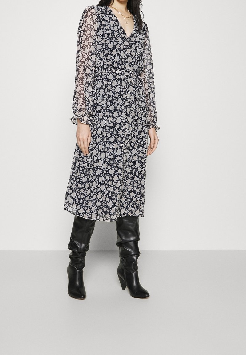 Femme portant une robe midi à fleurs noire et blanche avec des manches transparentes et des bottes en cuir noires montant jusqu'aux genoux, debout devant un fond blanc.