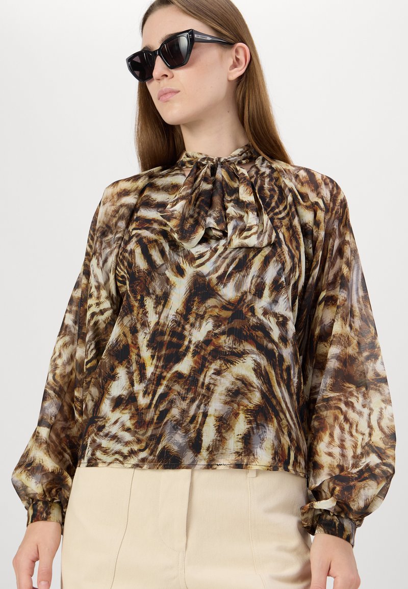 Transparente braune und beigefarbene Bluse mit einem Tigerstreifenmuster, hohem Kragen und langen Puffärmeln, kombiniert mit schwarzen Sonnenbrillen.