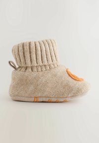 Botillon bébé beige tricoté avec revers pliable, visage souriant orange à l'avant et semelle antidérapante texturée sur fond uni.