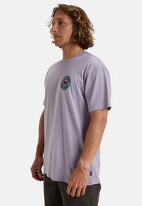 Camiseta de algodón de color púrpura claro con cuello redondo y mangas cortas, que presenta un logotipo gráfico circular en el lado izquierdo del pecho.