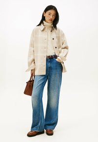 Beige plaid shirt jacket, cream knit turtleneck, wide-leg blue jeans, brown belt, and a small brown handbag. Flat brown loafers complete the look.