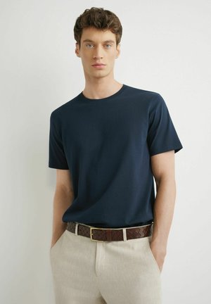 Młody mężczyzna z krótkimi kręconymi włosami, ubrany w granatowy t-shirt, beżowe spodnie i brązowy pleciony skórzany pasek, ręce w kieszeniach.