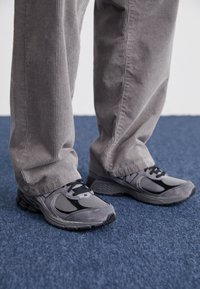 Šedé sportovní boty z prodyšného a semišového materiálu, s černými akcenty a tkaničkami. Nosené s šedými kordurovými kalhotami na modré podlaze.