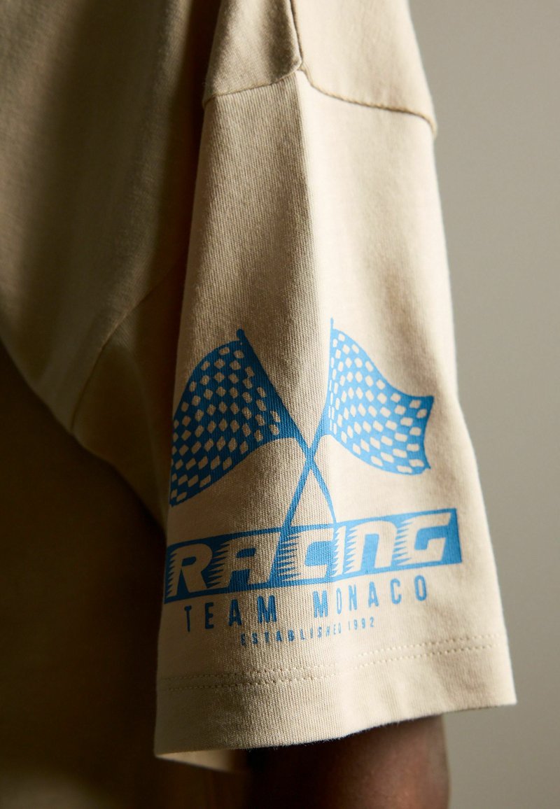 Beige Baumwoll-T-Shirt mit blauem Grafikdruck von zwei karierten Rennflaggen und dem Text "RACING TEAM MONACO" am Saum des Ärmels.