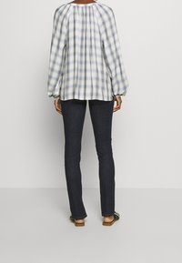 Blouse à carreaux bleu clair et blanc avec des manches volumineuses, un col froncé et une coupe décontractée, associée à un jean foncé et des sandales plates.