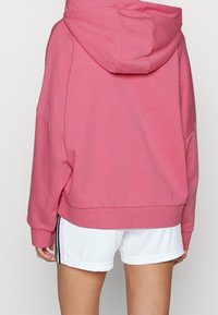 Felpa rosa con vestibilità ampia, dotata di un cappuccio spazioso e spalle scese. Abbinata a pantaloni corte bianchi con strisce laterali nere.