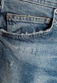 Modré džíny z denimu mají na zadní kapse opotřebovaný vzhled s třepícími se okraji a viditelnými švy, což naznačuje známky opotřebení s vybledlou texturou látky.