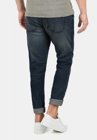 Muž v tmavě modrých jeansových kalhotách, které má vyhrnuté, a bílých teniskách, stojí s jednou nohou mírně zvednutou.