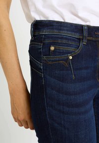 Vaqueros de mezclilla azul oscuro con un diseño entallado. Presentan costuras doradas, cuatro bolsillos y costuras laterales distintivas.