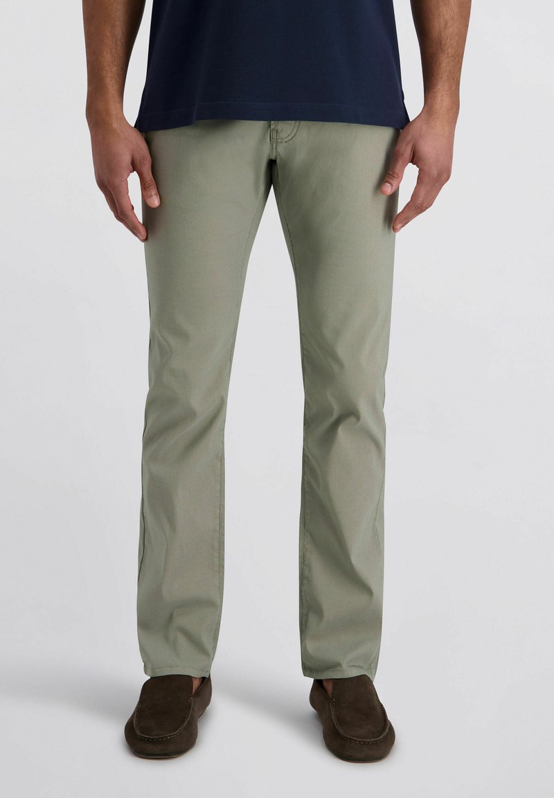 Mann trägt olivgrüne gerade Hose, dunkelbraune Loafers und ein dunkelblaues Hemd vor einem schlichten hellen Hintergrund.
